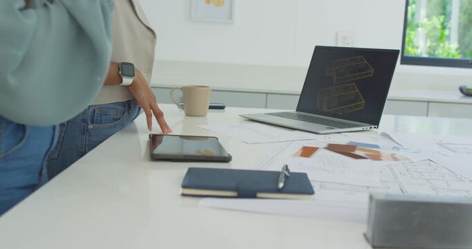 Adult woman reviewing plans, tapping tablet while editing design with laptop at counter, copy space