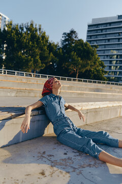 relaxed woman in denim jumpsuit and red headscarf lounging outdoors on concrete steps under bright sunlight with urban background and greenery