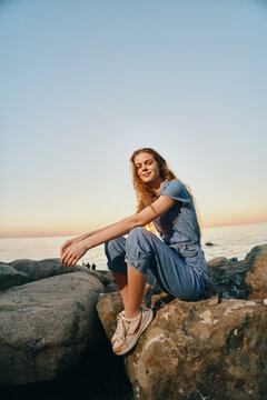 lifestyle woman in modern boho-western denim street style sitting on rocks by the sea during warm sunset with film color tones and relaxed vibe