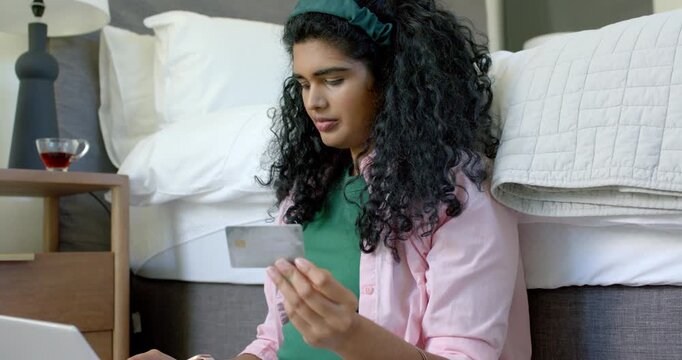 Woman holding payment card while typing on laptop at bedroom floor, smiling and confirming purchase