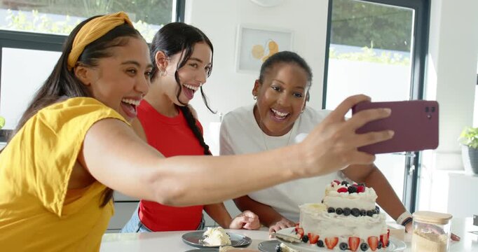 Diverse women raising purple phone taking selfies checking pics, celebrating cake at kitchen island
