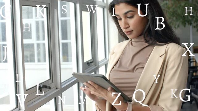 Woman standing by grid windows tapping tablet reading tech data, floating letters overlaying screen