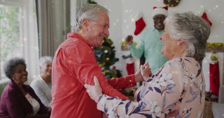 Naklejka premium Dancing senior couple wearing red shirt and floral blouse, holding hands in living room with tree
