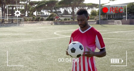 Naklejka premium Standing man wearing red-and-white kit, holding black-white ball on turf, showing REC 00:04:36