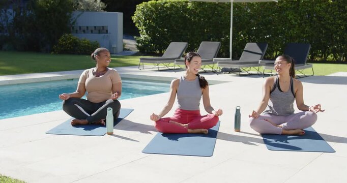 Three women sitting on blue mats poolside in leggings opening eyes center starting handhold bonding