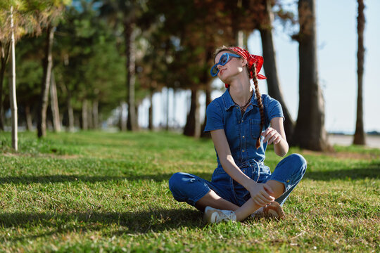 Lifestyle woman in modern boho-western denim street style sitting on grass outdoors, wearing sunglasses and red bandana, with film color effect in bright daylight.