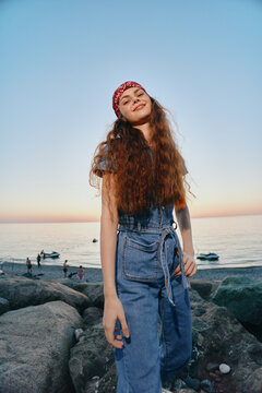smiling woman with red bandana wearing denim dress standing on rocky beach at sunset with calm sea and boats in background