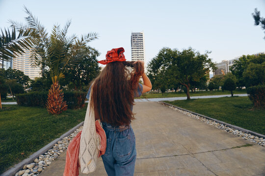 Lifestyle woman in modern boho-western denim street style walks outdoors with long hair, a red hat, and crochet sweater in film color tones on urban pathway.