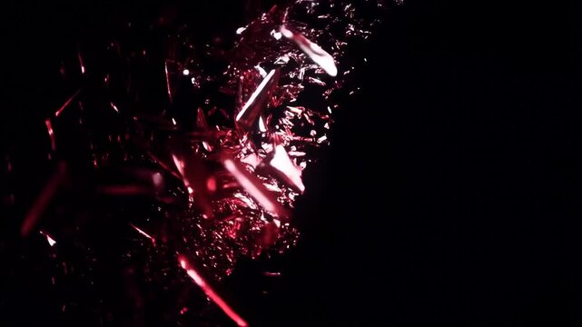 Slow motion tracking shot of red reflective glass shards exploding outward and scattering in closeup against black studio background