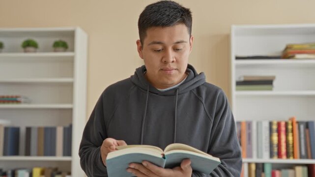 Man holding open book and turning a page amid bookshelves in a study building, focused on text; quiet concentration.