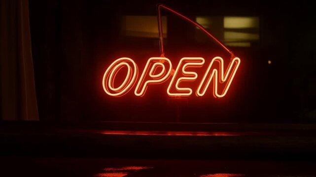 Tracking dolly slow zoom of neon open sign brightening with window reflection on wet pavement outside storefront at night