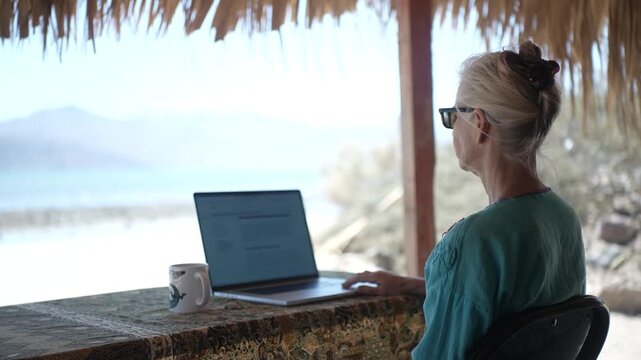 Mature woman sits at a table under a palapa working on her laptop while enjoying the beach view in Baja California, Mexico