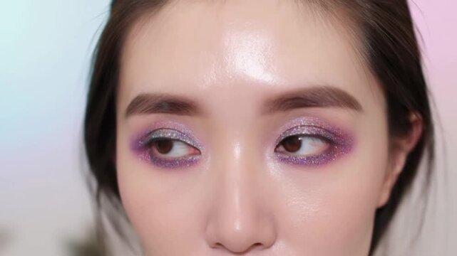 Closeup tracking shot of woman with purple glitter eye makeup shifting gaze and blinking across studio beauty tutorial set