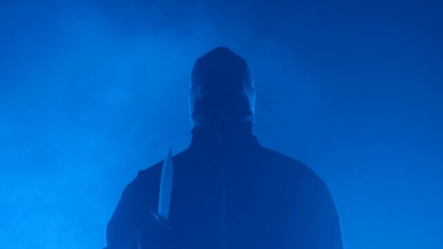 Static shot of masked figure walking toward camera through blue smoke holding knife in dark backlit studio