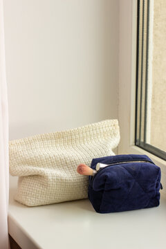 A windowsill scene with a cream woven cosmetic bag, a dark blue quilted makeup pouch with a brush.