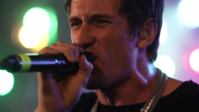 Handheld tracking shot of male singer gripping microphone and singing on concert stage with colorful bokeh lights at night