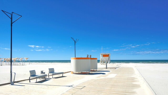 Modern empty beach with white sand, changing cabins, and wooden boardwalk under clear blue sky. Luxury resort infrastructure and seaside promenade. Summer vacation and travel concept.
