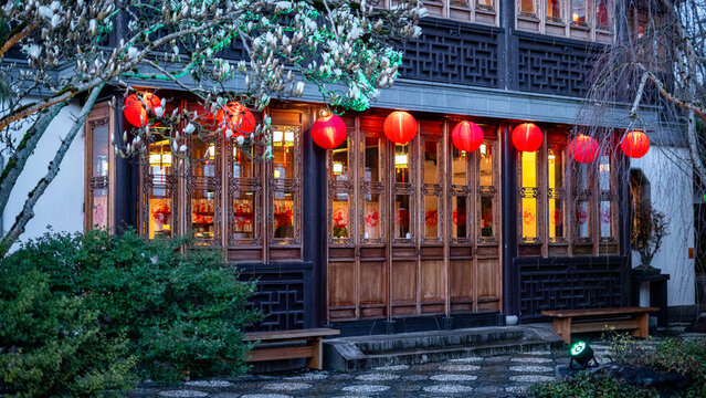 View of the traditional architecture of Lan Su Chinese Garden with red lanterns and blooming magnolia trees at dusk in Portland, Oregon, United States.
