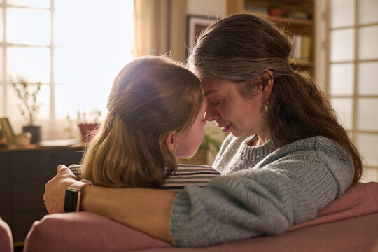 Caucasian mother and daughter embracing closely and touching foreheads on sofa, expressing comfort, trust, emotional support. Useful for parenting, family care, bonding, mental wellbeing themes
