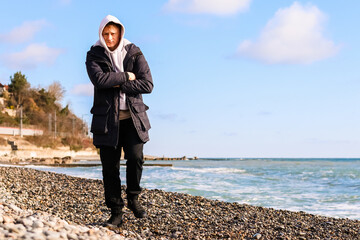 Man in hoodie stands arms crossed on pebble shore looking at sea © Michael