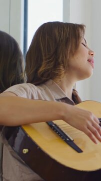 Vertical video: Adjusting hand singer in tan shirt strumming guitar and practicing song by window