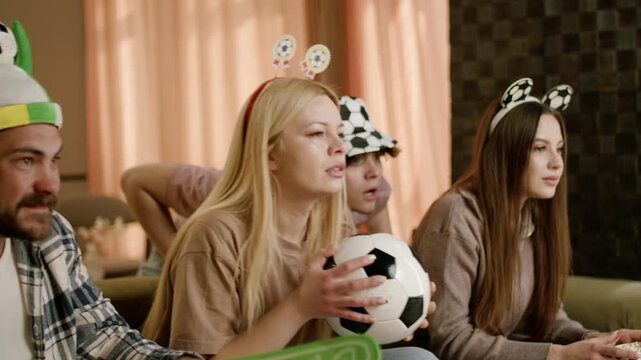 Group of friends wearing soccer-themed hats and accessories intensely watch a soccer match on television, holding a soccer ball and cheering in a cozy living room setting