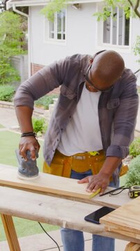 Vertical video: Picking up orbital sander, man sanding board on bench in front yard, smoothing wood