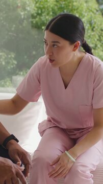 Vertical video: Talking nurse in scrubs guiding senior off couch at home holding watch helping up