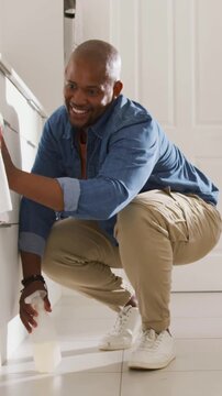 Vertical video: Placing bottle on tile, crouching man wiping white appliance in kitchen, cleaning