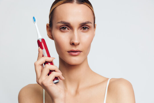 Young woman holds a red toothbrush near her face in a bright studio, focusing on oral care and dental hygiene, clean white background, self care and beauty routine for healthy teeth