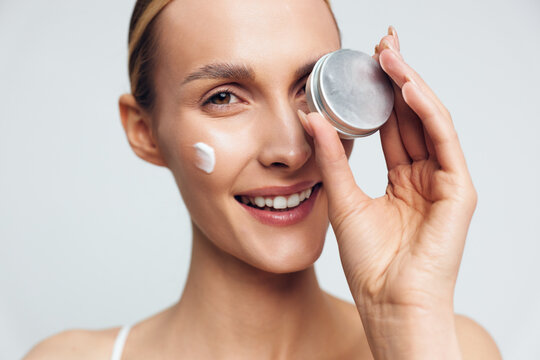 Smiling woman applying white face cream from a small jar. Closeup on a clean light background, focusing on skincare and selfcare beauty. Soft warm studio lighting.