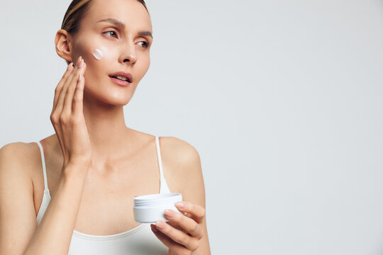 Young woman applies facial cream with fingertips, holding a skincare jar. Bright studio background highlights beauty care routine, selfcare, and healthy skin concept.