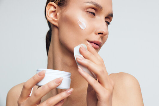 Woman skincare, face cream, applying moisturizer with closed eyes, holding jar and cotton pads, smooth skin, calm beauty routine, studio neutral background, daily care