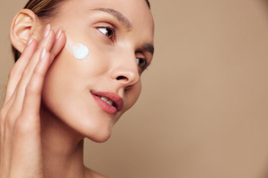 Woman applying skincare moisturizer to face in closeup beauty portrait, smooth skin, gentle hand touch, soft light, calm expression, beige clean studio background