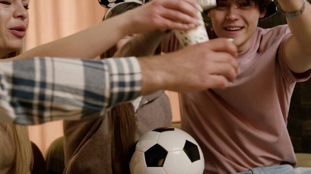 Group of young friends celebrating together while watching a soccer match, holding drinks and a soccer ball in a cozy indoor setting with festive decorations