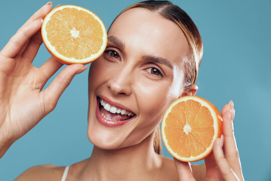 Happy woman holds orange slices near her face and smiles brightly, using citrus for skincare beauty concept, serum like glow, fresh fruit on blue background, copy space