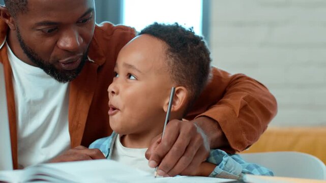 A Black man guides a young boy as he completes his homework. They sit at a table filled with books and learning materials, enjoying quality time at home.