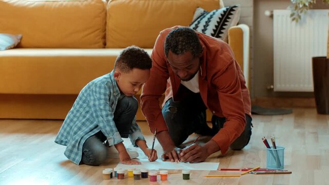 A man with dark skin and a boy are sitting on the floor of a living room. They are focused on drawing and painting on a sheet of paper. Art supplies are scattered around them.