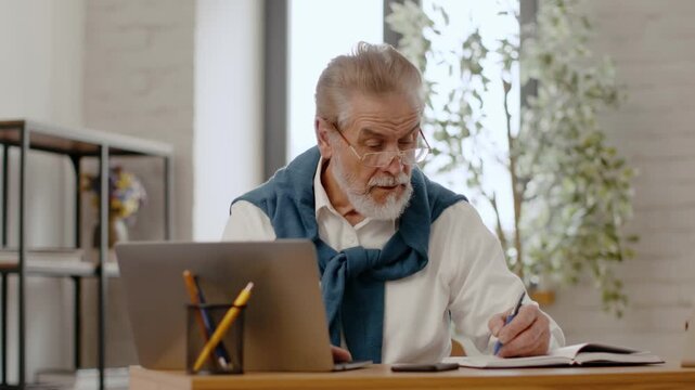 Senior man with a beard and glasses is sitting at a desk with a laptop. He appears focused while writing notes. The office has plants and modern decor.