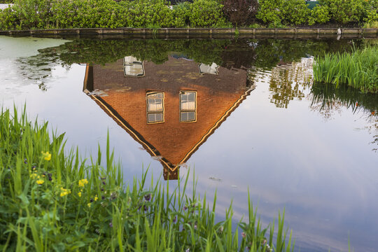 View of the reflection of a traditional brick house with a gabled roof in a calm canal surrounded by green grass and bushes Netherlands.