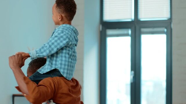 A father lifts his joyful son onto his shoulders inside their bright home. The boy wears a blue shirt and enjoys the fun. Windows allow natural light to fill the space.