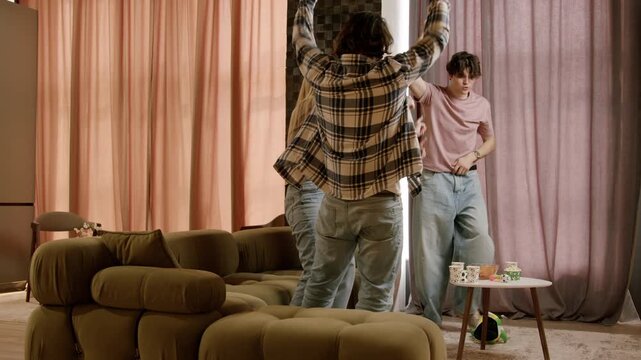 Group of friends dancing joyfully in a cozy living room with a green sofa, colorful curtains, and a small table with drinks and snacks visible in the background