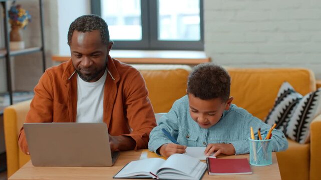A man with dark skin works on a laptop while sitting at a table. A boy with curly hair writes in a notebook beside him. They are in a well-lit living room filled with natural light.