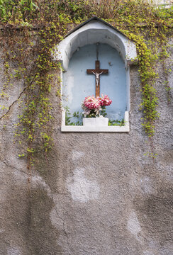View of a small religious shrine with a crucifix and pink flowers set into a weathered stone wall with hanging green vines in Italy.