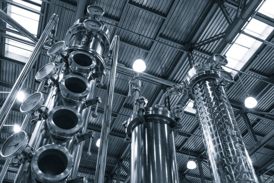 interior of an industrial distillery features tall copper and stainless steel columns. Pressure gauges and pipes indicate the ongoing distillation process. The high ceiling features exposed beams.