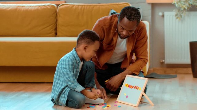 A man with dark skin sits on the floor with a young boy. They explore colorful letters and numbers together on a small easel. The cozy living room features a sofa and plants.