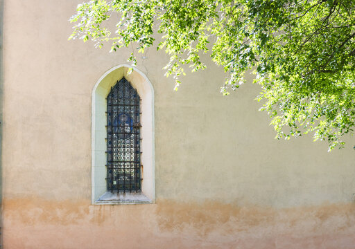 View of a pointed arch window with a metal grate on a textured stone wall partially shaded by green tree leaves Italy.