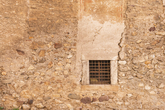 View of an ancient textured stone wall featuring a small square window with a rusty iron grate and weathered plaster Italy.