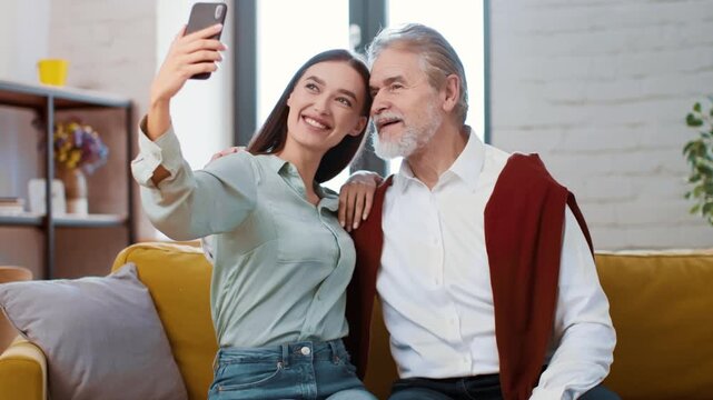 A young woman sits on a couch talking to an older man. They are engaged in conversation while the woman shows something on her phone. Natural light fills the room.