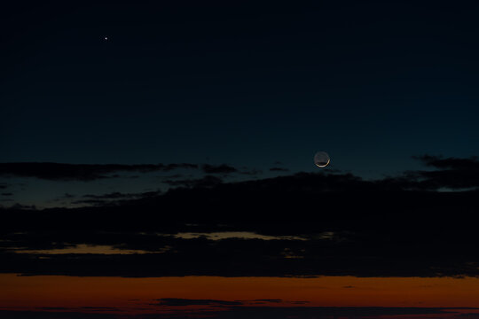View of a thin crescent moon and a bright star shining in the dark night sky above a horizon with orange sunset glow and dark clouds in Kyiv, Kyiv city, Ukraine.
